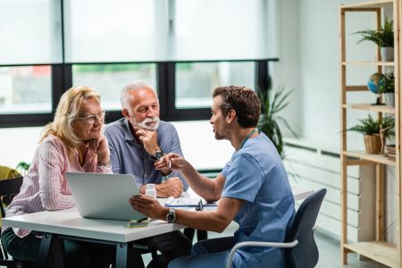 Happy mature couple and their doctor communicating while using laptop during medical appointment.